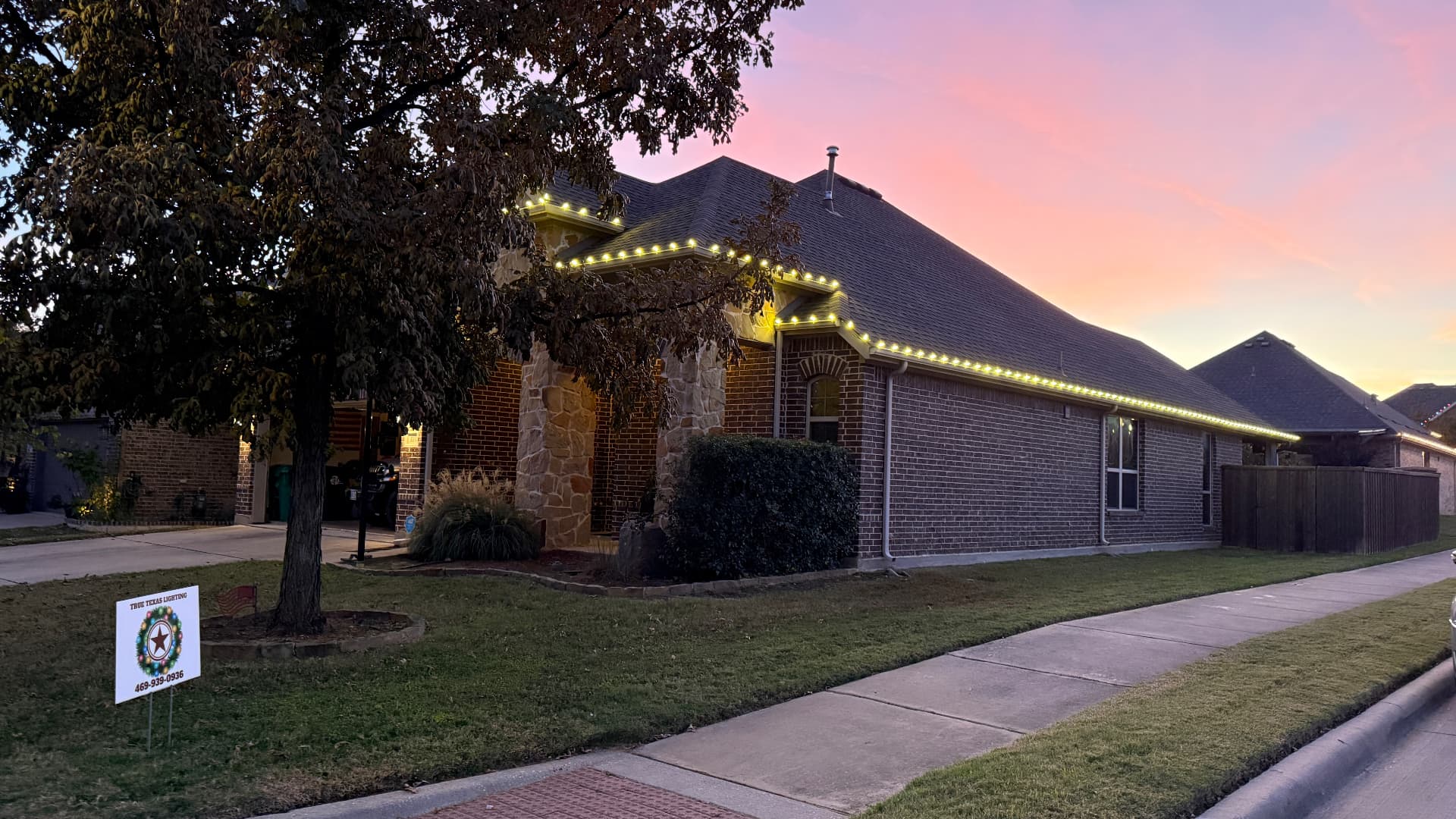Warm white roofline lights glowing at dusk with a True Texas Lighting yard sign